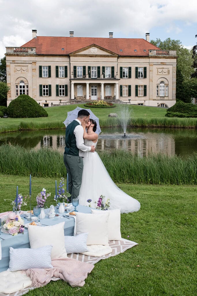 Hochzeitspaar küsst sich vor Schloss Harkotten mit Spitzenschirmchen, im Vordergrund gedeckter Picknicktisch